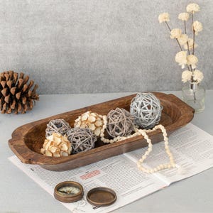 May include: A wooden decorative bowl filled with decorative spheres in various textures and colors, including woven, beaded, and floral designs. A compass and a dried flower arrangement are also present.