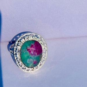 May include: Silver ring with an oval gemstone, displaying teal and magenta hues. The ring has a decorative, openwork border. The gemstone is the focal point of this piece of jewellery. A striking accessory.