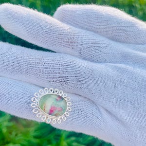 May include: Oval ring with a silver band and a light green and pink stone. The stone is set in a decorative silver setting with a scalloped edge. The ring is displayed on white fabric.