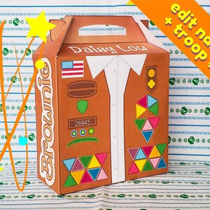 May include: A brown cardboard box with a Girl Scout Brownie troop logo. The box is decorated with a vest design, including a US flag, a badge, and a troop number 26945. The text "Daisy Lou" is printed on the box.