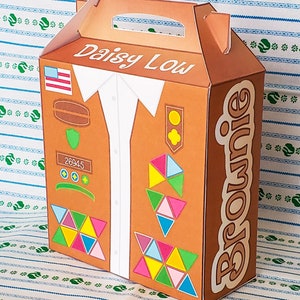 May include: A brown cardboard box with a handle, designed to look like a Girl Scout uniform. The box has the words "Daisy Low" and "Brownie" printed on it. The uniform features a white shirt, a brown vest with patches, and a green sash. The box is perfect for storing Girl Scout cookies.