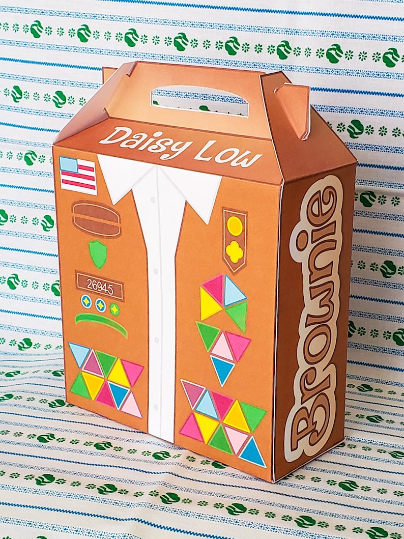 May include: A brown cardboard box with a handle, designed to look like a Girl Scout uniform. The box has the words "Daisy Low" and "Brownie" printed on it. The uniform features a white shirt, a brown vest with patches, and a green sash. The box is perfect for storing Girl Scout cookies.