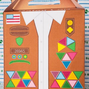 May include: A brown cardboard box with a cut-out handle. The box is decorated with a design of a Girl Scout uniform with a sash, badges, and patches. The text "Daisy Low" is printed on the box. The number "26945" is printed on the box.