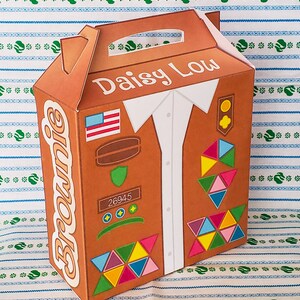 May include: A brown cardboard box with a handle, designed to look like a Girl Scout uniform. The box has the words "Daisy Low" and "Brownie" printed on it, along with a patch that says "26945".
