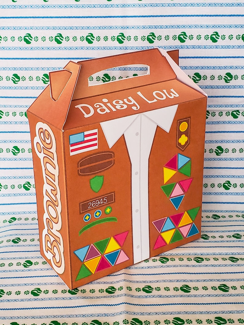 May include: A brown cardboard box with a handle, designed to look like a Girl Scout uniform. The box has the words "Daisy Low" and "Brownie" printed on it, along with a patch that says "26945".