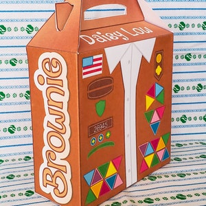 May include: A brown cardboard box with a handle, decorated with a Girl Scout uniform design. The box has the words "Brownie" and "Daisy Lou" printed on it.