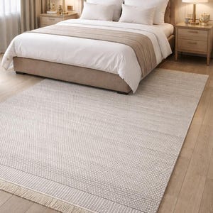 Puede incluir: Una alfombra de dormitorio de tonos neutros con textura tejida y bordes con flecos. La alfombra es de color gris claro y beige, y está colocada frente a una cama con ropa de cama blanca y un cabecero beige. El ambiente es cálido.