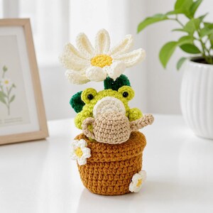 Può includere: Figurina di rana all'uncinetto fatta a mano con un grande fiore di margherita, foglie verdi e un annaffiatoio. La rana è in un vaso marrone con fiori bianchi e gialli. Un delizioso oggetto decorativo.