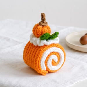 Op de afbeelding: Een gehaakte oranje en witte Zwitserse rol als decoratie, bekroond met een kleine oranje pompoen, een bruine steel en twee groene blaadjes. De cake heeft een spiraalpatroon van oranje en wit garen met een witte gehaakte rand aan de bovenkant.