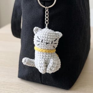 Puede incluir: Un llavero de gato tejido a ganchillo en color gris claro con un collar amarillo y detalles faciales negros bordados, incluyendo ojos y bigotes. La figura del gato está unida a una anilla de metal plateado y una cadena corta, colgada de un bolso de tela negro.