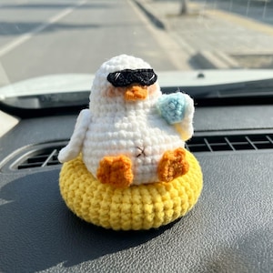 Peut inclure: Une figurine de caneton crochetée en blanc portant des lunettes de soleil noires et tenant un petit objet bleu clair. Le caneton a des pattes et un bec orange, et il est assis dans un anneau crocheté jaune vif rappelant une bouée.