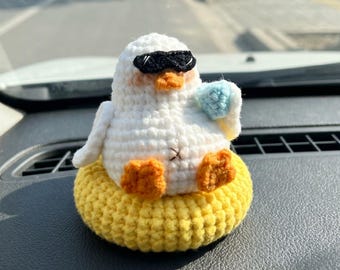 May include: A white crocheted duckling figure wearing black sunglasses and holding a small light blue object. The duckling has orange feet and a beak, and it is sitting inside a bright yellow crocheted ring that resembles a pool floatie.