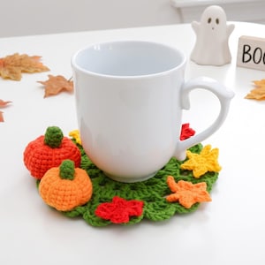May include: A plain white ceramic mug sits on a round green crocheted coaster decorated with two small orange pumpkins and several colorful autumn leaves in red, orange, and yellow. The coaster features a textured, open-work yarn design.