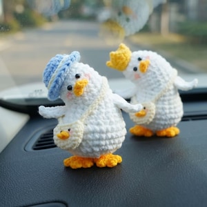 May include: Two small, white, crocheted duck figurines. The front duck wears a light blue hat and carries a small cream-coloured bag. The second duck behind it wears a yellow crown and also carries a small cream-coloured bag. Both have orange feet and beaks.