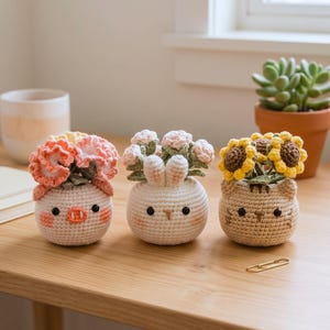 Puede incluir: Tres macetas de ganchillo hechas a mano con forma de cerdo, conejo y gato, cada una con flores de ganchillo de colores. La maceta de cerdo tiene flores melocotón y coral, el conejo tiene flores rosas y el gato tiene girasoles amarillos.