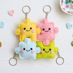 Puede incluir: Cuatro llaveros de piezas de rompecabezas de ganchillo de colores amarillo, rosa, azul y verde, cada uno con una cara sonriente y mejillas rosadas. Los llaveros están dispuestos juntos, con llaveros de metal.