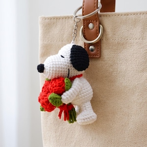 May include: A white and black crocheted dog keychain holding a bouquet of red flowers with green stems. The keychain is attached to a silver metal ring and chain, which is clipped onto the brown leather strap of a beige canvas tote bag.