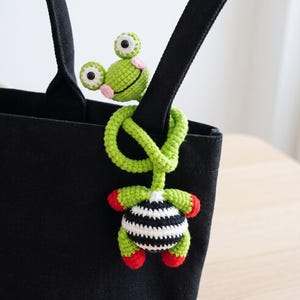 May include: A green crocheted frog figure with large white eyes, pink cheeks, and a long flexible body wrapped around the strap of a black canvas tote bag. The frog has a round black and white striped belly and red accents on its hands and feet.