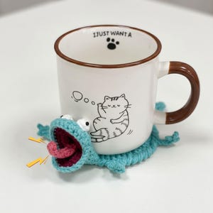 Op de afbeelding: Een witte keramische mok met een bruine rand en oor, voorzien van een zwarte lijntekening van een kat en de tekst I JUST WANT A met een pootafdruk. De mok staat op een lichtblauwe gehaakte onderzetter in de vorm van een kikker met open mond.