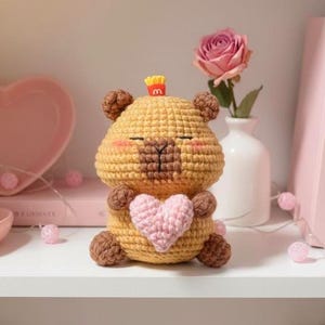 May include: A handmade, crocheted bear figurine in shades of tan and brown, holding a pink heart. The bear has a small french fry charm on its head. The figurine is displayed on a white shelf with a pink rose and other decorative items.