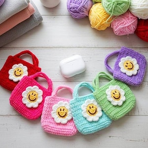 Könnte beinhalten: Eine Sammlung bunter, gehäkelter Mini-Tragetaschen, jede mit einem lächelnden Gänseblümchen-Applikation verziert. Die Taschen sind in verschiedenen Farbtönen erhältlich, darunter Rot, Rosa, Blau und Grün. Wollknäuel in passenden Farben und ein weißes Etui sind ebenfalls vorhanden.