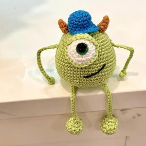 Peut inclure: Une poupée monstre ronde en crochet vert clair avec un seul grand œil blanc, un sourire cousu noir, deux petites cornes brunes et une casquette de baseball en crochet bleu vif. La poupée a des bras et des jambes fins avec des pieds ronds.