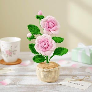 Può includere: Una composizione floreale all'uncinetto fatta a mano con due rose rosa, foglie verdi e un bocciolo, in un vaso beige all'uncinetto. La composizione è su una superficie di legno bianco, con una tazza e una confezione regalo sullo sfondo. Una carta dice "Mother's Day".