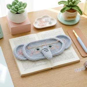 May include: A crocheted elephant-shaped glasses holder in light gray, with pink accents and green eyes, rests on an open book. Gold-rimmed glasses sit on the holder. The scene includes crocheted plants, a cloud-shaped dish, and a pen.