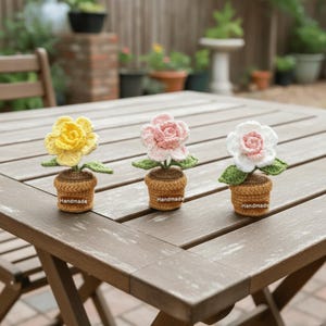 May include: Three handmade crochet flowers in small pots sit on a wooden table. The flowers are yellow, pink, and white, each with green leaves. The pots are brown with "Handmade" labels.