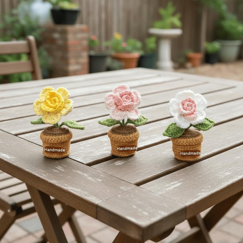 May include: Three handmade crochet flowers in small pots sit on a wooden table. The flowers are yellow, pink, and white, each with green leaves. The pots are brown with "Handmade" labels.