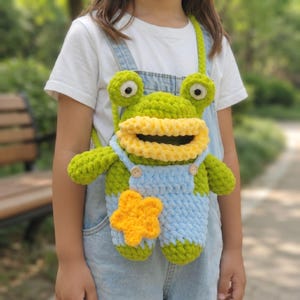 May include: A handmade crochet frog bag featuring a bright green body, large eyes, and a wide yellow mouth. The frog wears light blue crocheted overalls with two small wooden buttons and a prominent yellow star appliqué on the front panel.