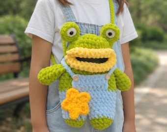 Könnte beinhalten: Eine handgefertigte Häkeltasche in Froschform mit leuchtend grünem Körper, großen Augen und einem breiten gelben Mund. Der Frosch trägt eine hellblaue Häkel-Latzhose mit zwei kleinen Holzknöpfen und einem gelben Stern-Applikations-Motiv.