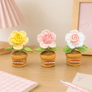 May include: Three handmade crochet flower arrangements. Each flower is a different color: yellow, pink, and white. The flowers sit in small brown pots with the word "Handmade" on them. Green leaves are visible.
