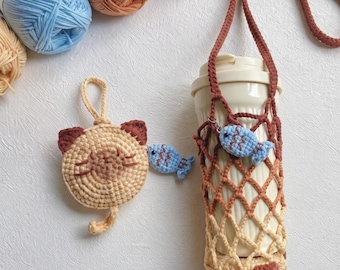 Könnte beinhalten: Ein beiges, gehäkeltes Täschchen in Katzenform mit braunen Ohren und Schnurrhaaren sowie einem kleinen blauen Fisch-Anhänger. Daneben steht ein cremefarbener Kunststoffbecher in einem braun-beigen, gehäkelten Netzhalter mit langem Trageriemen.
