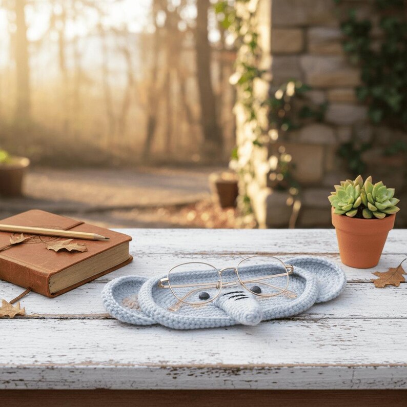 May include: A light blue, crocheted elephant-shaped glasses holder with gold-rimmed glasses resting on it. A brown leather-bound journal with a pencil and a small potted succulent are also on the white wooden surface. The background is blurred.