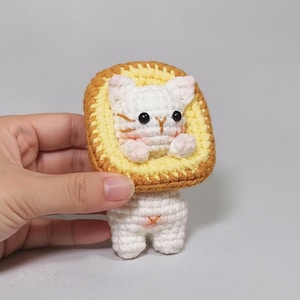 Puede incluir: Un juguete de gato hecho a mano, tejido a ganchillo, con un cuerpo blanco y una cabeza en forma de pan amarillo y marrón. El gato tiene ojos negros, mejillas rosas y pequeñas patas. El juguete está sobre un fondo gris claro.