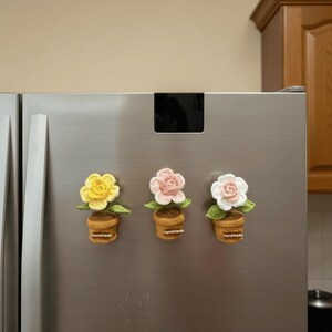 May include: Three handmade crochet flower magnets in yellow, pink, and white, each in a brown pot with green leaves. The word "Handmade" is on each pot. The magnets are attached to a stainless steel refrigerator door.