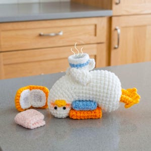 Op de afbeelding: Een gehaakte witte eendenfiguur met oranje voeten en snavel, met een klein blauw-wit kopje op de rug met witte stoomlijntjes erboven. Naast de eend liggen een klein oranje-wit rechthoekig stukje en een gevouwen lichtroze vierkantje.