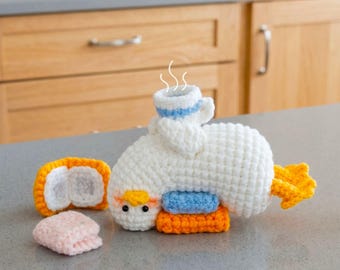 May include: A white crocheted duck figure with orange feet and beak, featuring a small blue and white cup on its back with white steam lines above it. Beside the duck are a small orange and white rectangular piece and a folded light pink square.