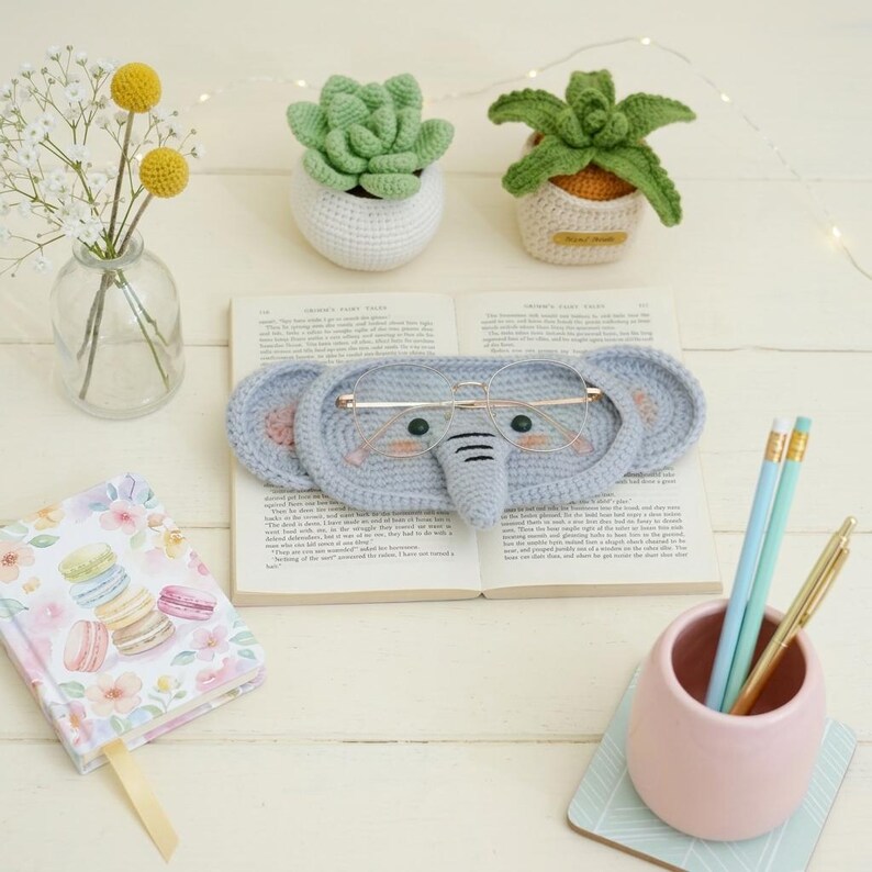 May include: A light blue crocheted elephant-shaped glasses holder with a trunk and pink ears, holding a pair of glasses. The holder rests on an open book, with two crocheted succulent plants and a small notebook with a floral design.