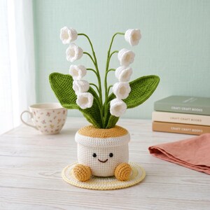 May include: A crocheted lily of the valley plant in a white and yellow pot with a smiling face. The plant features white bell-shaped flowers, green leaves, and yellow crocheted feet. The pot sits on a yellow crocheted coaster.