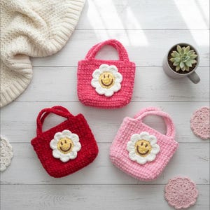 K&ouml;nnte beinhalten: Drei handgefertigte geh&auml;kelte Tragetaschen in Rot, Rosa und Ros&eacute;, jede mit einer wei&szlig;en G&auml;nsebl&uuml;mchen-Applikation mit gelbem Smiley-Gesicht. Die Taschen haben passende Griffe und sind auf einer hellen Holzoberfl&auml;che ausgestellt.