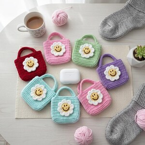 K&ouml;nnte beinhalten: Eine Sammlung farbenfroher, geh&auml;kelter Mini-Tragetaschen, jede mit einem l&auml;chelnden G&auml;nsebl&uuml;mchen-Gesicht verziert. Die Taschen sind in verschiedenen Farben erh&auml;ltlich, darunter Rot, Rosa, Gr&uuml;n, Blau und Lila. Ein wei&szlig;es AirPod-Etui liegt neben den Taschen, zusammen mit einer Kaffeetasse, einer Sukkulente und grauen Socken.
