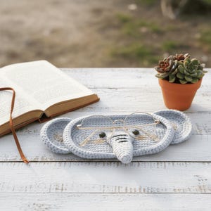 May include: A light blue crocheted elephant-shaped glasses holder with gold-rimmed glasses resting on it. The holder has two round ear-shaped sections and a trunk. A small potted succulent and an open book are in the background.