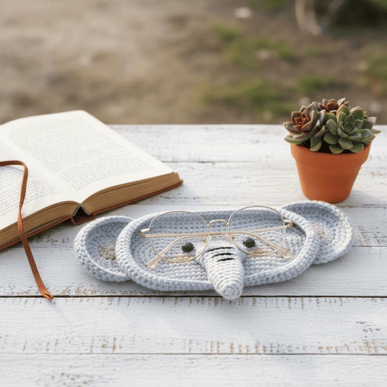 May include: A light blue crocheted elephant-shaped glasses holder with gold-rimmed glasses resting on it. The holder has two round ear-shaped sections and a trunk. A small potted succulent and an open book are in the background.