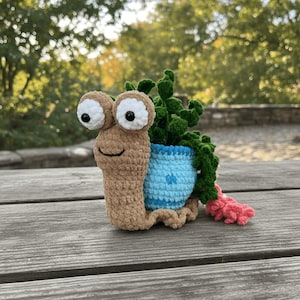 Puede incluir: Una maceta de caracol de ganchillo hecha a mano. El caracol es de color marrón claro con grandes ojos saltones y una cara sonriente. Contiene una maceta azul con una planta verde y una cola rosa. La maceta está sobre una superficie de madera.