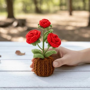 Può includere: Un'arrangiamento floreale all'uncinetto fatto a mano con tre rose rosse brillanti, foglie e steli verdi, presentato in un piccolo vaso marrone. L'immagine mette in risalto la dettagliata trama del filato e l'arte del design.