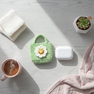 K&ouml;nnte beinhalten: Eine Draufsicht mit einer hellgr&uuml;nen H&auml;keltasche mit einem wei&szlig;en G&auml;nsebl&uuml;mchengesicht, einem wei&szlig;en Etui, einer Tasse Kaffee, einer kleinen Sukkulente in einem wei&szlig;en Topf und zwei wei&szlig;en Waschlappen. Eine rosa Decke ist auf der rechten Seite des Bildes drapiert.