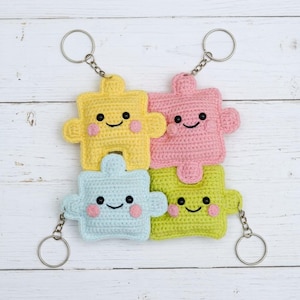 May include: Four crocheted puzzle piece keychains in yellow, pink, light blue, and green. Each piece has a smiling face with rosy cheeks and is attached to a silver keyring. The keychains are arranged in a square formation.