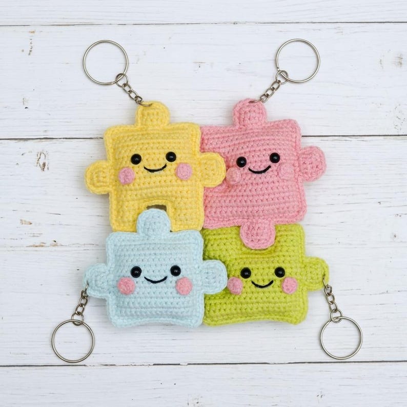 May include: Four crocheted puzzle piece keychains in yellow, pink, light blue, and green. Each piece has a smiling face with rosy cheeks and is attached to a silver keyring. The keychains are arranged in a square formation.